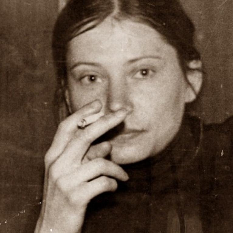 A sepia-toned photograph of a young woman with dark hair pulled back, holding a cigarette near her face. The photo captures a close-up portrait with a neutral expression and a plain background.
