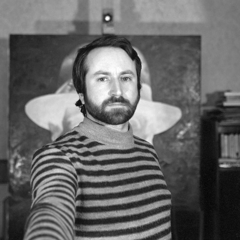 Photograph of a man with a beard and mustache wearing a striped turtleneck sweater, taking a selfie. Background includes a large painting of a figure with a hat and a bookshelf filled with books.