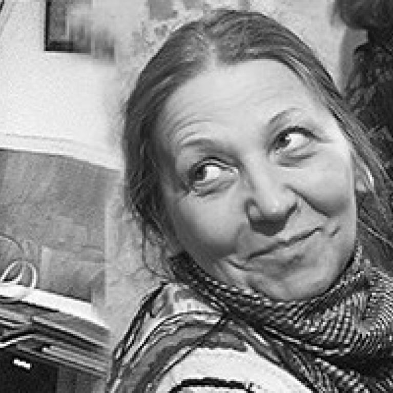 Black and white photograph showing a woman with a playful expression looking sideways, wearing a patterned scarf. Background includes framed pictures on a wall, a vase with flowers, and other objects.