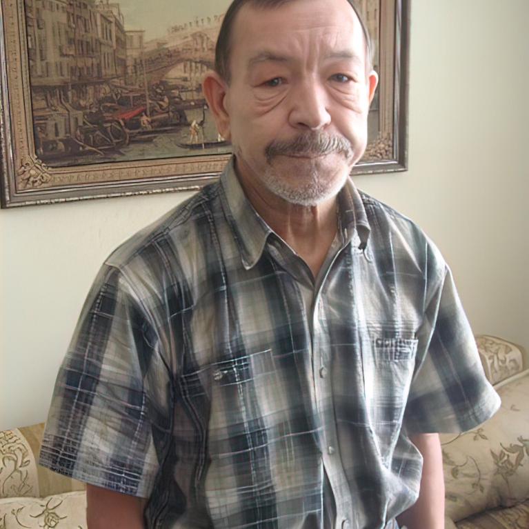 Photograph of an older man standing indoors in front of a couch and a framed painting depicting a canal scene. The man wears a short-sleeve plaid shirt tucked into blue jeans with a belt, and has a neutral expression with hands in pockets.