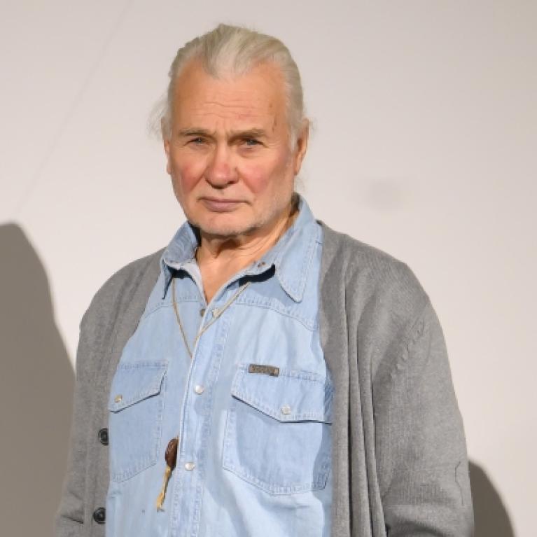 Photograph of an older man with white hair pulled back, wearing a light blue denim shirt under a gray cardigan. He stands against a plain light background, looking directly at the camera with a neutral expression.
