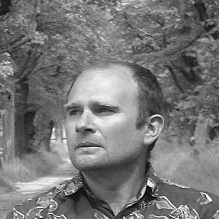 Black and white photograph of a man with a receding hairline wearing a patterned shirt, standing on a tree-lined path. The background features dense foliage creating a natural tunnel effect, emphasizing a contemplative mood.