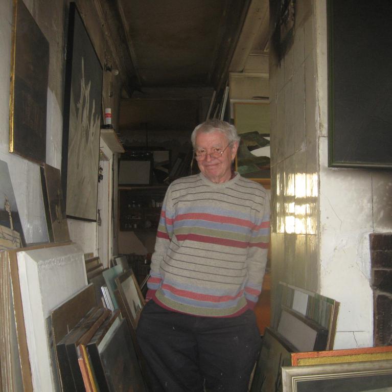 Photograph of an elderly man standing in a narrow room filled with stacked framed paintings and artworks leaning against walls. The man wears a striped sweater and glasses, smiling gently, with a fireplace visible on the right and dim lighting creating a cozy, cluttered atmosphere.