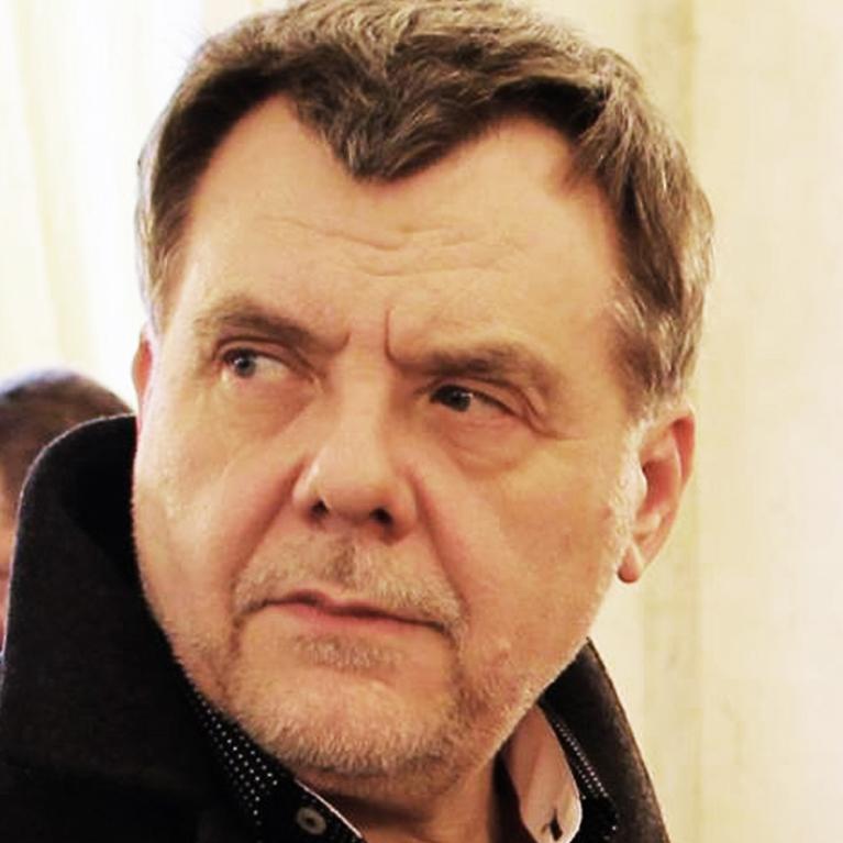Photograph of a middle-aged man with short brown hair and a trimmed beard, wearing a black coat and a dark shirt with a subtle pattern. The man is looking slightly to his left with a neutral expression, set against a softly lit, neutral-colored background.