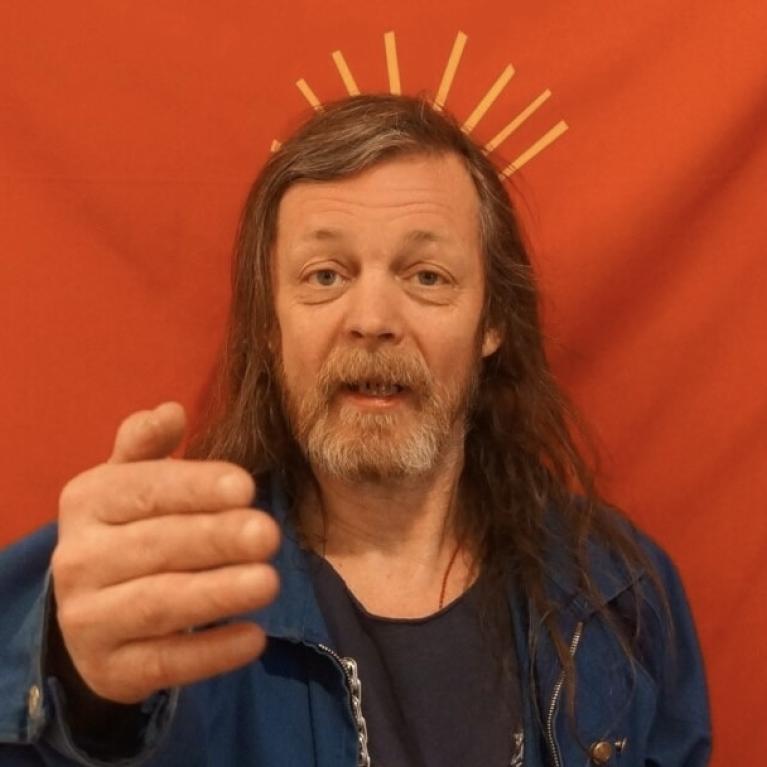 Photograph of a man with long hair and a beard wearing a blue jacket, standing against a red background. Yellow lines behind his head create a halo effect, giving a symbolic or artistic impression.