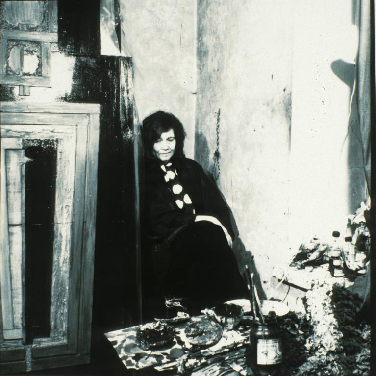 Black and white photograph of a woman sitting on the floor in a corner of a room, surrounded by art supplies including paint jars and brushes. The setting suggests an artist's studio with textured walls and a wooden cabinet, highlighting a moment of rest or contemplation.