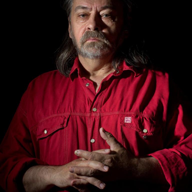 Photograph of a middle-aged man with long hair and a beard wearing a red button-up shirt, standing against a black background. The lighting highlights his face and hands, creating a dramatic and contemplative mood.