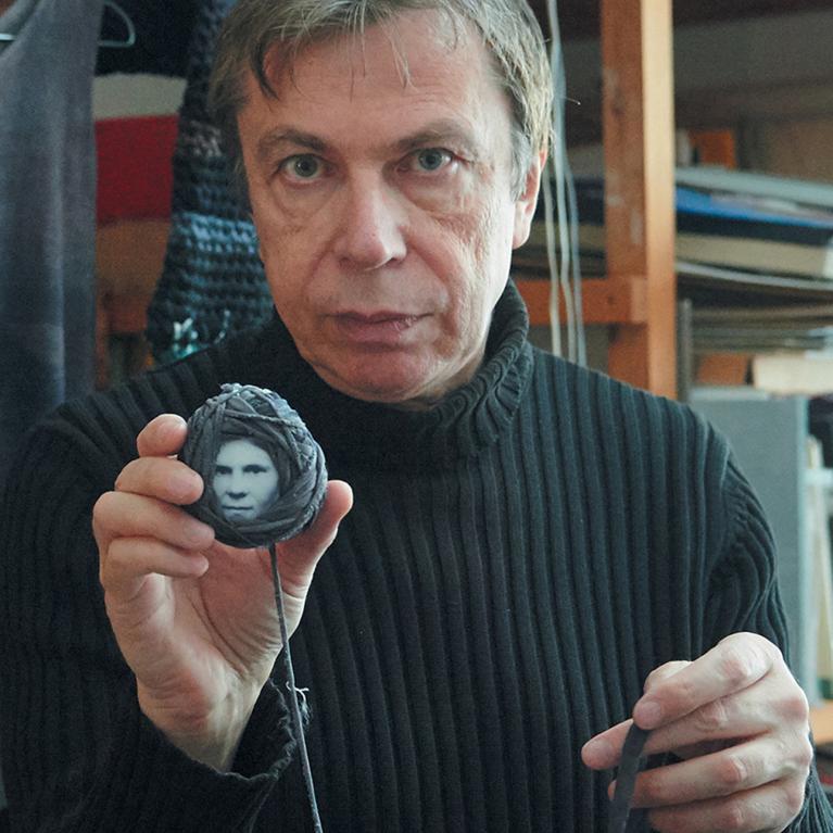 Photograph of a man wearing a black ribbed sweater holding a small circular object with a black-and-white face image wrapped in dark thread. Background shows shelves with books and other items, suggesting a creative or work environment.