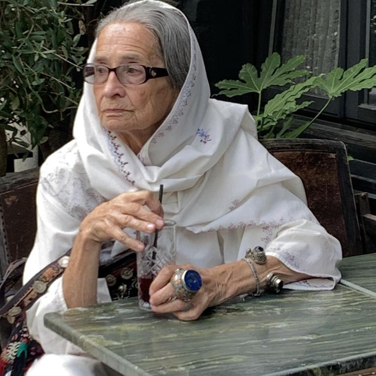 Photograph of an elderly woman wearing glasses and a white shawl, sitting at a green marble table outdoors. She holds a glass with a straw and wears multiple rings and bracelets, with a contemplative expression and greenery in the background.