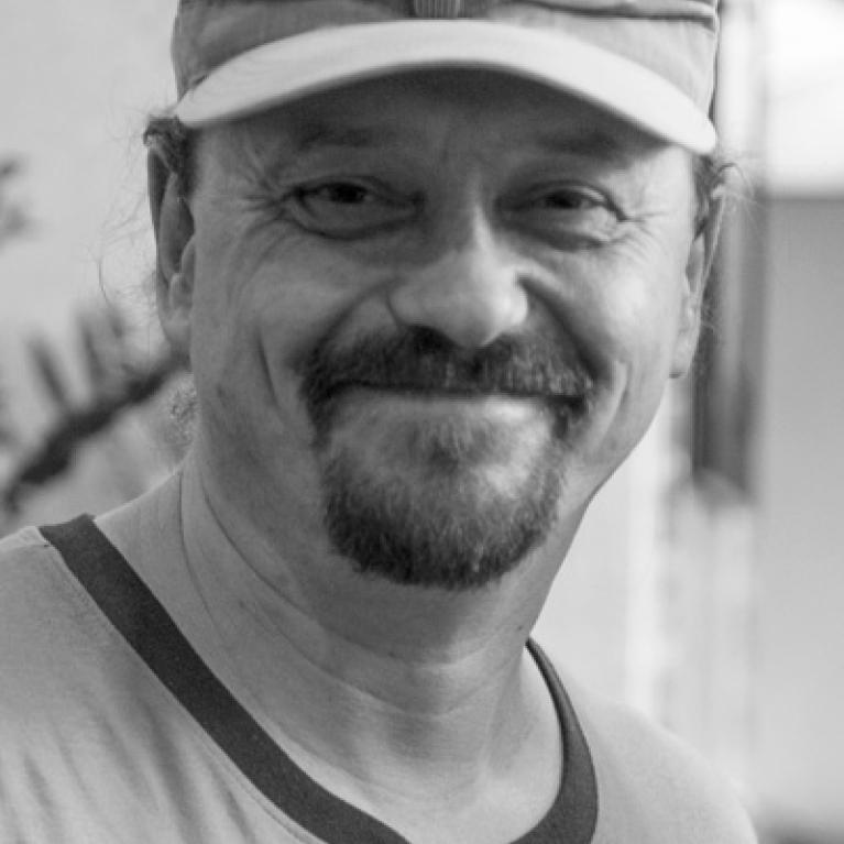 Black and white photograph of a smiling man wearing a light-colored cap and a t-shirt with a small pin on the left side. The background is blurred, focusing attention on the man's facial expression and casual attire.