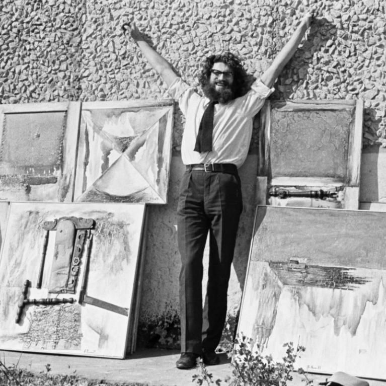 Black and white photograph of a man standing outdoors with arms raised, surrounded by five abstract paintings leaning against a textured wall. The man wears glasses, a tie, and dark pants, and the paintings feature geometric shapes and textured brushstrokes in varying shades of gray.