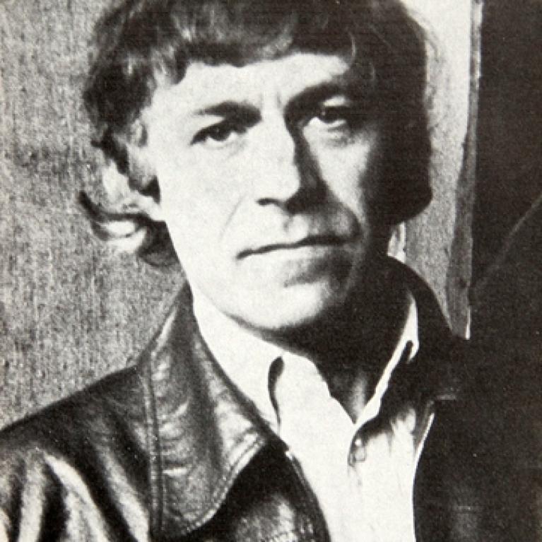 Black and white photograph of a man with medium-length hair wearing a leather jacket over a collared shirt. 