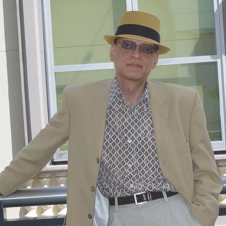 Photograph of a man wearing a tan blazer, patterned shirt, beige pants, and a tan fedora with a black band, standing casually with one arm resting on a railing. Background features large windows and a light-colored wall, suggesting an outdoor or balcony setting.