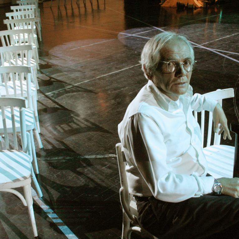 Photograph of an elderly man with glasses sitting on a white wooden chair in a dimly lit gymnasium or auditorium with a row of identical white chairs extending behind him. 