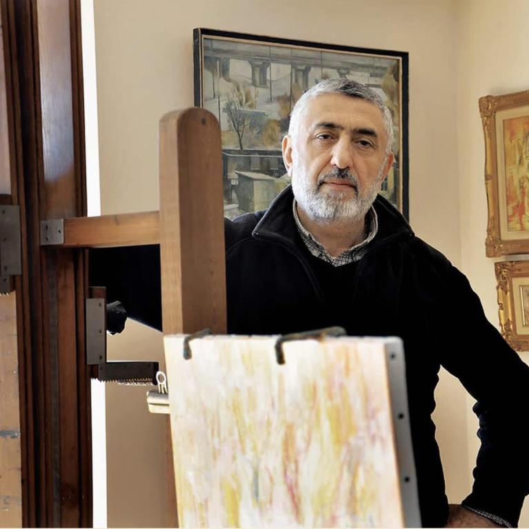 Photograph of a man standing beside an easel with a canvas in a room decorated with framed paintings. The man, wearing a black jacket, appears to be an artist in a studio or gallery setting, with the canvas showing an abstract or unfinished artwork.