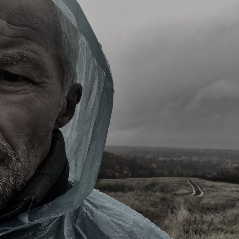 Photograph of a man wearing a rain poncho standing outdoors on a cloudy, overcast day. Background shows a winding dirt path leading through a grassy field toward a distant tree line under a gray sky.