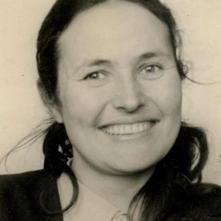 Black and white photograph of a smiling woman with long dark hair pulled back, wearing a collared shirt and dark jacket. The photo captures a close-up portrait with a neutral background, highlighting her friendly expression.
