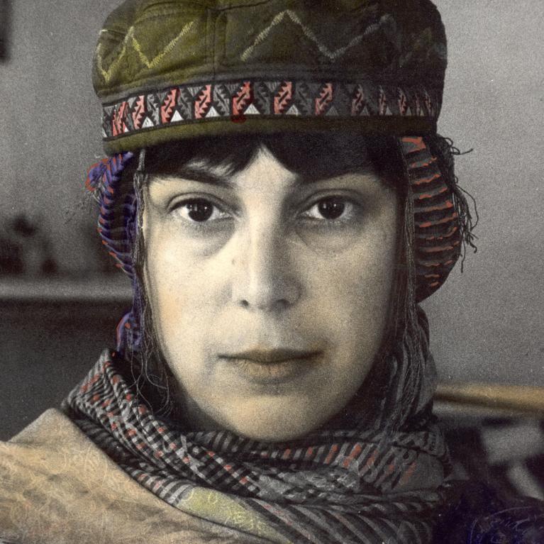 Photograph of a woman wearing a patterned scarf and a green cap with decorative trim, looking directly at the camera. 