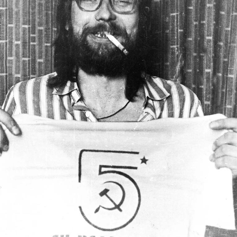 Black-and-white photograph shows bearded, long-haired man wearing glasses and smoking a cigarette while holding a T-shirt. Shirt features stylized numeral five, hammer-and-sickle emblem, star, and Cyrillic lettering, suggesting provocative political or countercultural messaging.