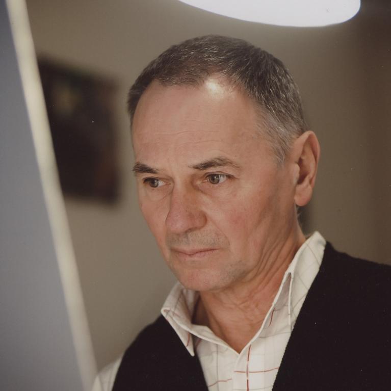 Portrait photograph of a middle-aged man wearing a black sweater over a white, red-lined shirt indoors. Warm ceiling light and blurred background frame his neutral, attentive expression.