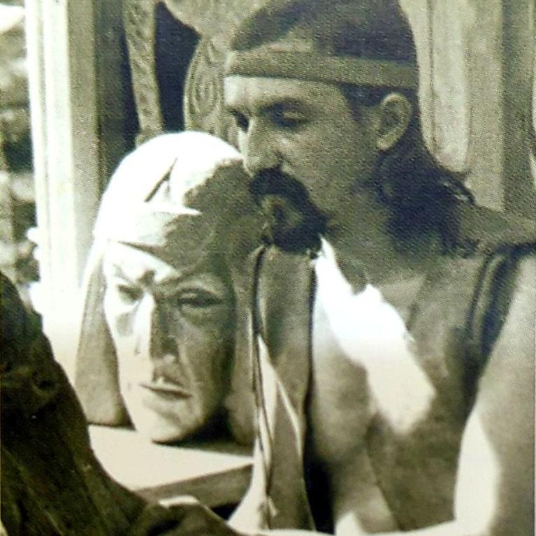 Black-and-white photograph shows a shirtless, long-haired man holding a pale human-head prop beside a table or sculpture. 