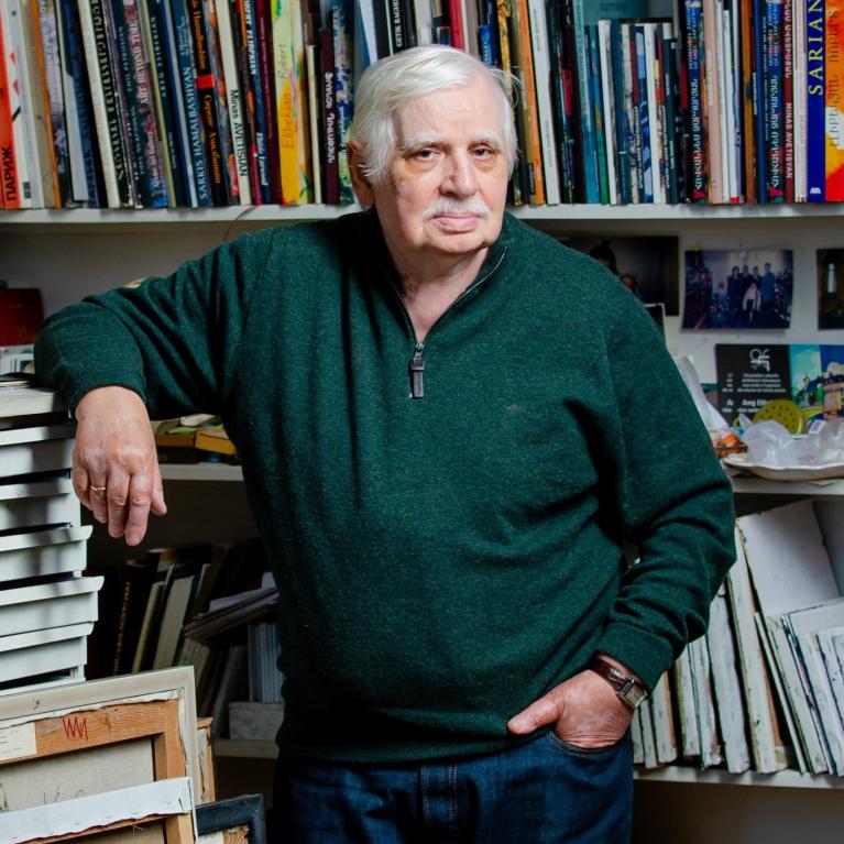 photo portrait of a grey-haired man with moustache in green sweater in front of bookshelves