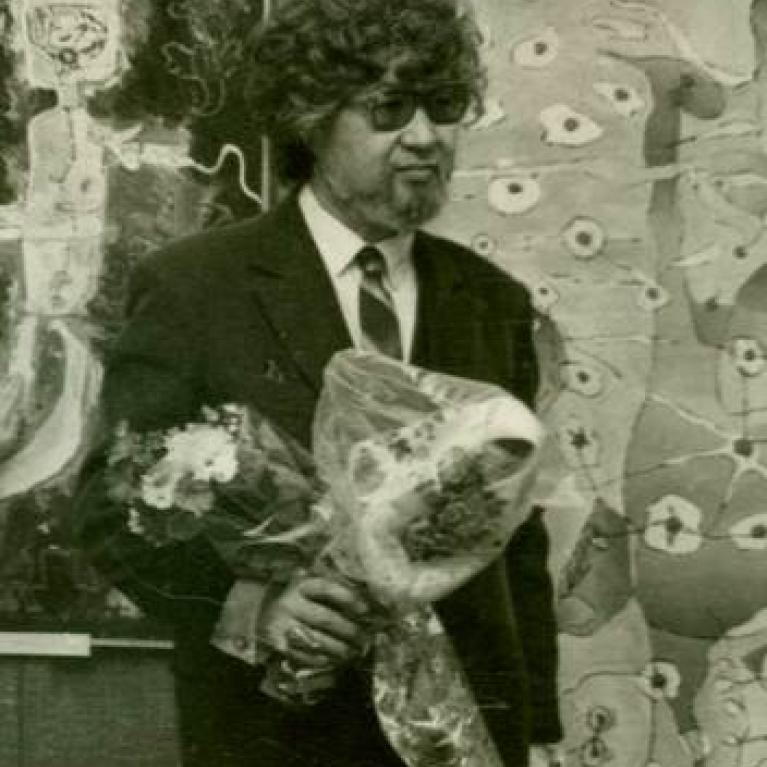 Black-and-white photograph shows a man in a suit and sunglasses holding a large flower bouquet inside an art gallery. 