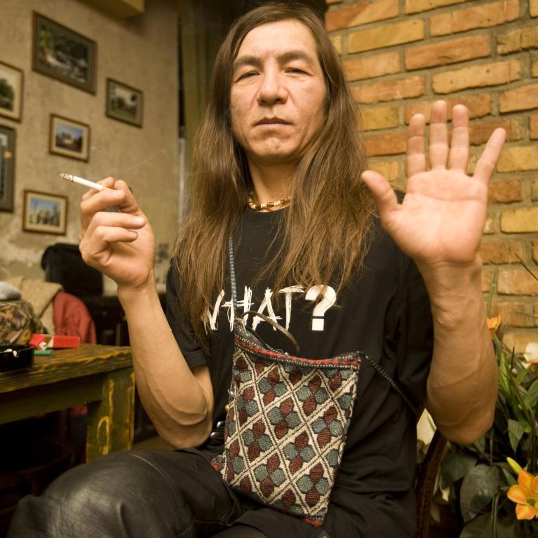 Photograph of long-haired man seated indoors beside exposed brick, holding a cigarette and raising one hand toward camera. Framed pictures, arched doorway, plants, and black shirt marked “WHAT?” provide room context.