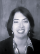 A black-and-white photograph of a person wearing a dark jacket and patterned shirt standing against a wooden wall background. 