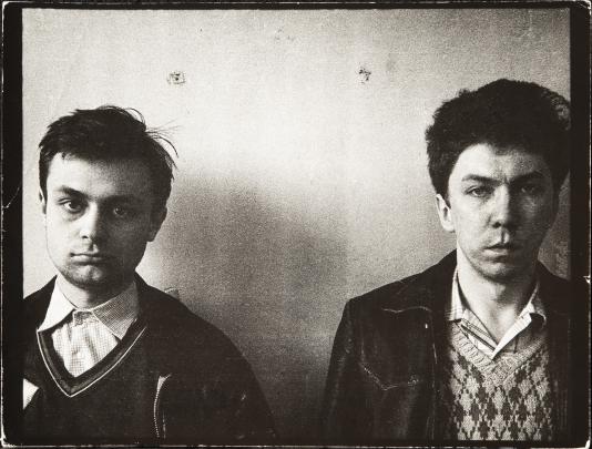 Black and white photograph showing two young men standing side by side against a plain wall. Both wear collared shirts under sweaters and jackets, and the photo has a vintage, worn border effect.