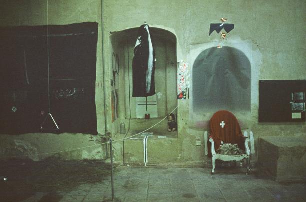 Photograph of a dimly lit, worn indoor space featuring a hanging fish in an arched alcove and a white chair draped with a red cloth marked by a white cross. Surrounding elements include a dark rectangular panel on the left, a small display on the right, and various wires and objects scattered on a tiled floor, evoking a mysterious or artistic atmosphere.