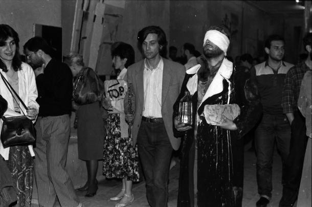 Black and white photograph showing a group of people standing in a hallway, with one man prominently blindfolded and wearing a long coat while holding a bottle and a loaf of bread.