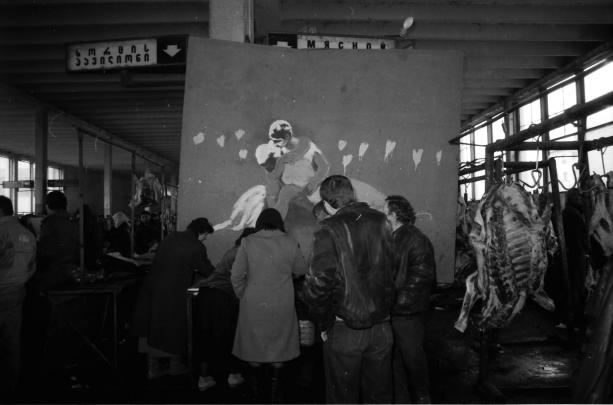 Black and white photograph of a group of people standing and working inside a meat processing facility with hanging carcasses on the right side. The scene includes a large wall with a faded painted figure, industrial ceiling beams, and signage in a non-Latin script above.