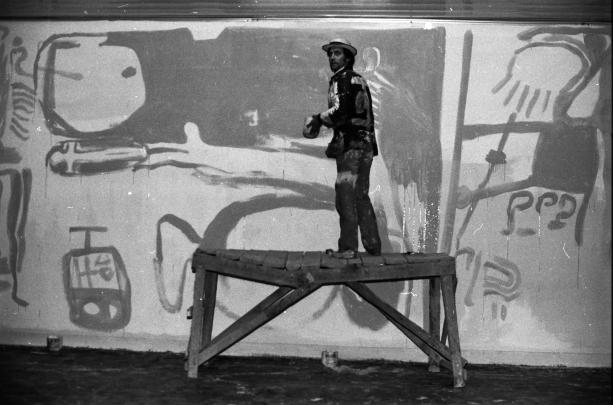 Black and white photograph showing a man standing on a wooden scaffold while painting a large mural on a wall. The mural features abstract shapes and figures, including a large face and various geometric patterns, highlighting an artistic process in progress.