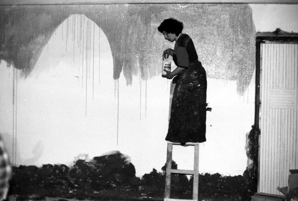 Black and white photograph showing a woman standing on a ladder painting a wall with a paint can in hand. Dark paint covers lower wall area and drips down from partially painted upper section, with a closed door visible on the right side.