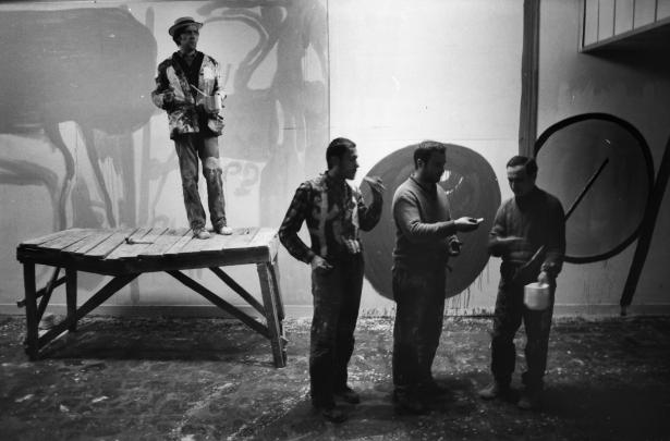 Black and white photograph showing four men in a studio setting, with one man standing on a wooden platform holding a paint bucket and brush, while three others stand on the floor engaged in conversation. Background features large abstract shapes painted on a wall, suggesting an artistic or creative environment.